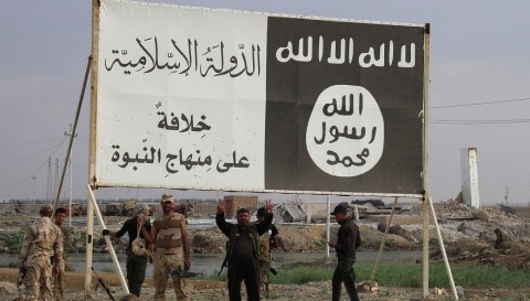 Iraqi government forces gather under a billboard bearing slogans of the Islamic State (IS) group and its trademark flag, in the town of Heet, in Iraq's Anbar province, which they are battling to retake from IS jihadists, on April 7, 2016. Earlier in the month Iraqi security forces recaptured parts of Heet, which was one of the largest population centres in Anbar province still held by the Islamic State (IS) group, but other areas remain under jihadist control. / AFP PHOTO / MOADH AL-DULAIMI