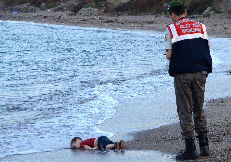 GRAPHIC CONTENT A Turkish police officer stands next to a migrant child's dead body (Aylan Shenu) off the shores in Bodrum, southern Turkey, on September 2, 2015 after a boat carrying refugees sank while reaching the Greek island of Kos. Thousands of refugees and migrants arrived in Athens on September 2, as Greek ministers held talks on the crisis, with Europe struggling to cope with the huge influx fleeing war and repression in the Middle East and Africa. (Photo by Nilufer Demir / DOGAN NEWS AGENCY / AFP)